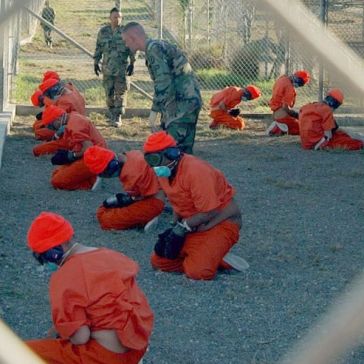 “Detainees in orange jumpsuits sit in a holding area under the watchful eyes of Military Police at Camp X-Ray at Naval Base Guantanamo Bay, Cuba, during in-processing to the temporary detention facility on Jan. 11, 2002. The detainees will be given a basic physical exam by a doctor, to include a chest x-ray and blood samples drawn to assess their health. DoD photo by Petty Officer 1st class Shane T. McCoy, U.S. Navy.”